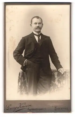 Fotografie Georg Knirim, Hannover, Portrait stattlicher Herr in Anzugjacke mit Krawatte an Tisch gelehnt