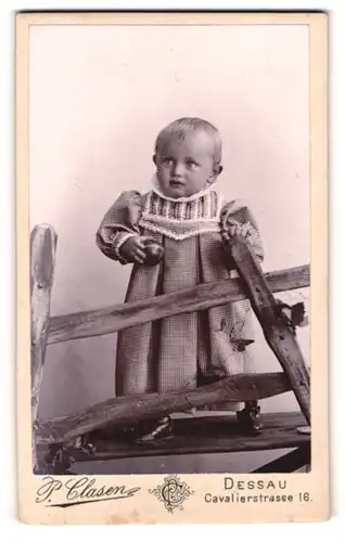 Fotografie P. Clasen, Dessau, Portrait süsses Kleinkind im karierten Kleid mit Ball an Zaun gelehnt