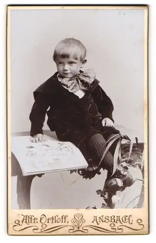 Fotografie Alfr. Ortloff, Ansbach, Portrait kleiner Junge im Samtanzug mit Fliege und Bilderbuch auf Tisch sitzend