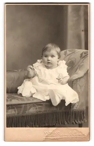 Fotografie Wolffram & Comp., Bremen, Baby in weissem Kleid sitzt auf bestickter Ottomane