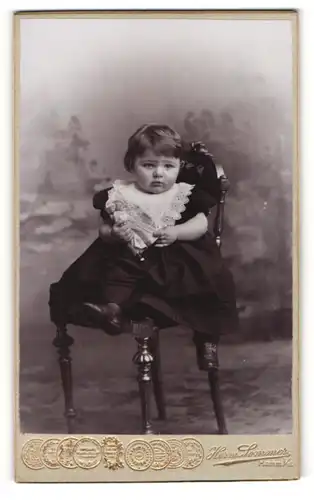 Fotografie Herm. Sommer, Hamm i / W., Portrait sitzendes Kleinkind im hübschen Kleid mit Puppe