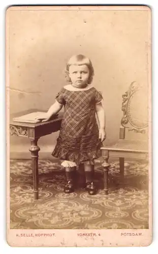 Fotografie Hermann Selle, Potsdam, Portrait kleines Mädchen im karierten Kleid mit Buch an Tisch gelehnt