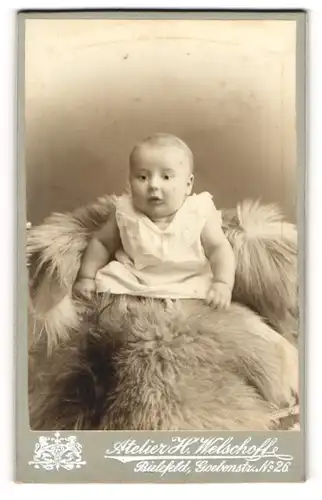 Fotografie Atelier H. Welschoffe, Bielefeld, Baby in weissem Laibchen sitzt auf Fell