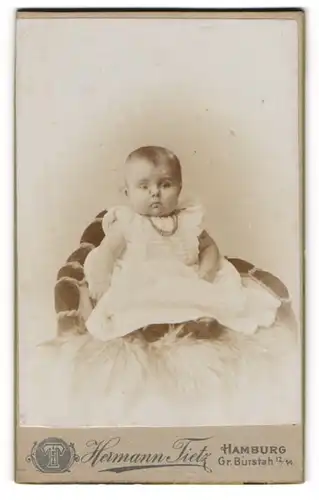 Fotografie Hermann Tietz, Hamburg, Kleinkind in weissem Kleidchen trägt Halskette und sitzt auf Fell