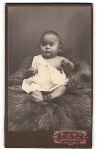 Fotografie Th. Dimmers, Hilden / Rhld., Portrait süsses Kleinkind im weissen Hemd auf Fell sitzend