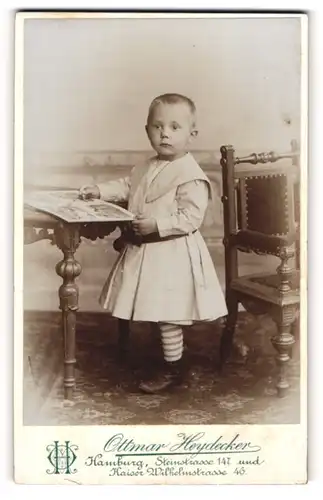 Fotografie Ottmar Heydecker, Hamburg, Kleiner Junge in weissem Kleid steht vor einem Tisch