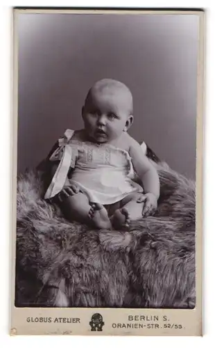 Fotografie Globus Atelier, Berlin, Baby im weissen Kleidchen auf Felldecke sitzend