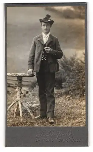 Fotografie H. Gutjahr, Ramsau /Bay., Junger Mann mit Trachtenhut und Zigarette vor Almlandschaft