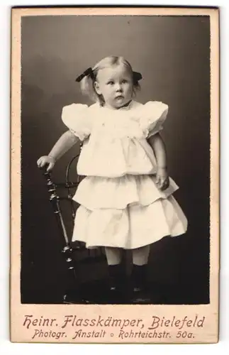 Fotografie Heinr. Flasskämper, Bielefeld, Rohrteichstr. 50, Niedliches Mädchen in weissem Kleid mit schwarzen Schleifen