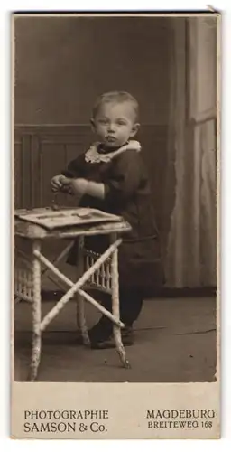 Fotografie Samson & Co., Magdeburg, niedliches blondes Kleinkind mit Buch am Tisch stehend