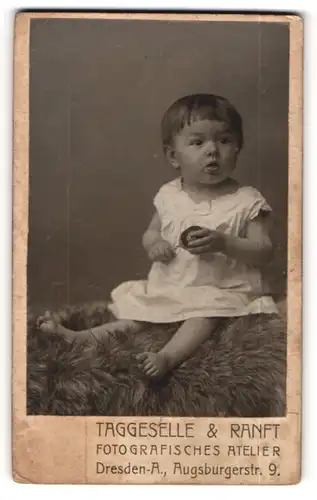 Fotografie Taggeselle & Ranft, Dresden-A., Portrait brünettes kleines Mädchen mit Rassel im weissen Kleidchen