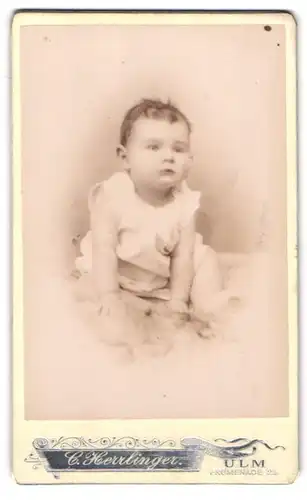 Fotografie C. Herrlinger, Ulm, Portrait sitzendes Kleinkind im weissen Hemd mit nackigen Füssen