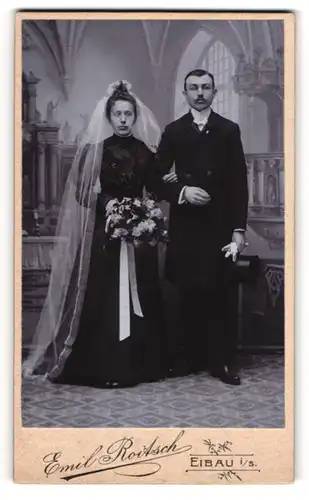 Fotografie Emil Roitsch, Eibau i. S., Brautleute im schwarzen Hochzeitskleid und Anzug vor einer Studiokulisse