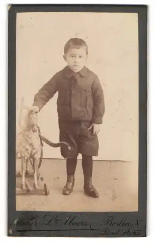 Fotografie L. Maerz, Berlin, Bad-Str. 65, Junger Bursche in schwarzen Klamotten mit Spielzeugpferd schaut gelangweilt