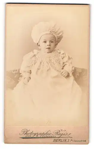 Fotografie Regis, Berlin, niedliches Kleinkind im weissen Kleid mit Mütze