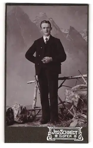 Fotografie Kos. Schmidt, Lofer, junger Mann im dunklen Anzug vor einer Studiokulisse