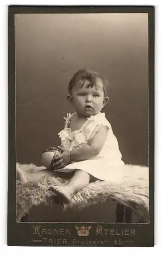 Fotografie Kronen-Atelier, Trier, Portrait niedliches Kleinkind im weissen Hemd mit Ball auf Fell sitzend