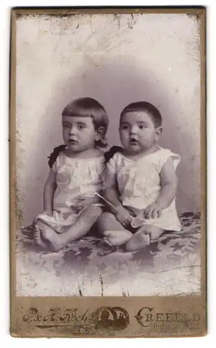 Fotografie P. & H. Koch, Crefeld, Portrait zwei niedliche Kleinkinder in weissen Hemden auf Tisch sitzend