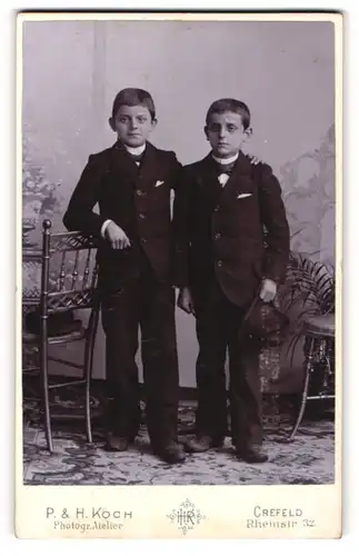 Fotografie P. & H. Koch, Crefeld, Portrait zwei kleine Jungen in Anzügen an Stuhl gelehnt