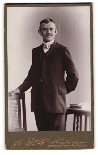 Fotografie H. Imig, Zwickau i / T., Portrait junger Herr im Anzug mit Fliege an Stuhl gelehnt