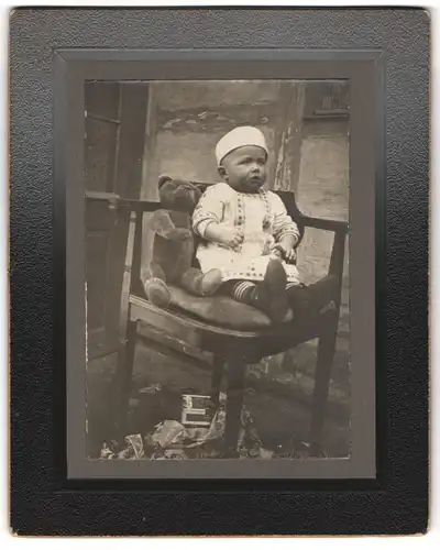 Fotografie unbekannter Fotograf und Ort, Kleinkind mit seinem Teddybär auf einem Stuhl sitzend