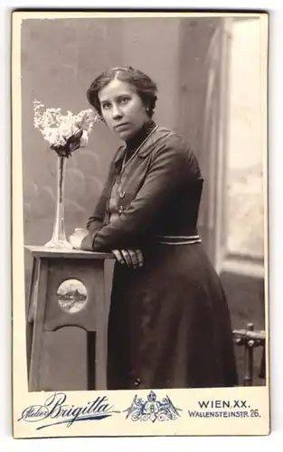 Fotografie Atelier Brigitta, Wien, Wallensteinstr. 26, Junge Dame in schwarzem Kleid lehnt an Tischchen mit Blumenvase