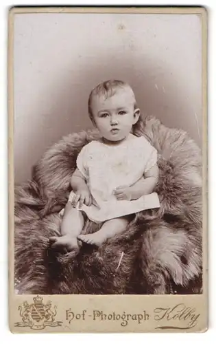 Fotografie Kolby, Chemnitz, Portrait niedliches Kleinkind im weissen Hemd auf Fell sitzend
