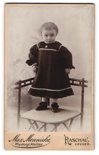 Fotografie Max Mennicke, Raschau / Erzgeb., Portrait niedliches Kleinkind im hübschen Kleid auf Stuhl stehend