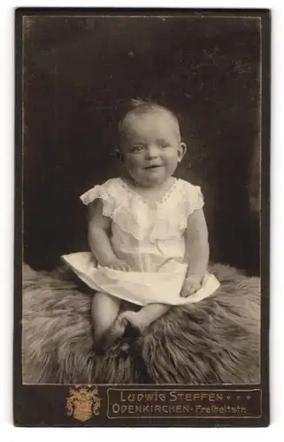 Fotografie Ludwig Steffen, Odenkirchen, lachendes süsses Kleinkind im weissen Kleidchen auf Fell sitzend