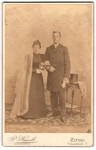 Fotografie P. Heinelt, Zittau, Ehepaar im schwarzen Brautkleid und Anzug, Zylinder auf Beistelltisch
