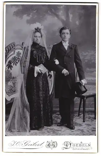 Fotografie J. O. Geilert, Döbeln, junges Ehepaar im schwarzen Brautkleid und Anzug mit Zylinder
