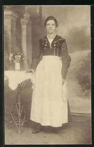 Foto-AK Junge Dame in Tracht mit Schürze an Tisch gelehnt vor Studiokulisse