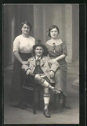 Foto-AK Sitzender Mann in Tracht mit zwei jungen Frauen vor einer Studiokulisse