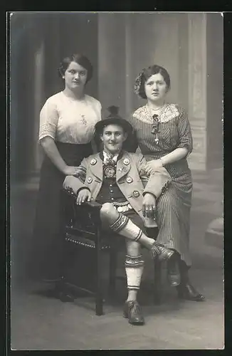 Foto-AK Sitzender Mann in Tracht mit zwei jungen Frauen vor einer Studiokulisse