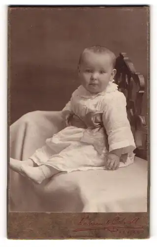 Fotografie Larsen & Ödvall, Arvika, Portrait niedliches Kleinkind im weissen Kleid auf Decke sitzend