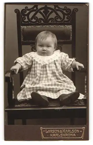 Fotografie Larson & Karlson, Karlskrona, Portrait niedliches Kleinkind im karierten Kleid auf Stuhl sitzend