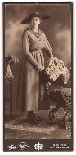Fotografie Max Fischer, Berlin-N, Portrait modisch gekleidete Dame mit Hut an Stuhl gelehnt
