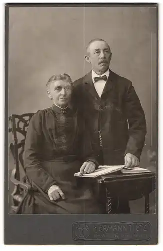 Fotografie Hermann Tietz, Hamburg, Portrait älteres Ehepaar in eleganter Kleidung mit Buch am Tisch