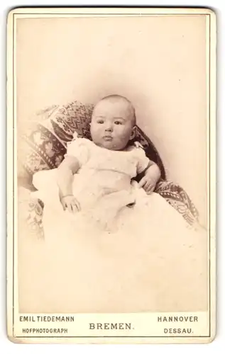 Fotografie Emil Tiedemann, Bremen, Portrait niedliches Baby im weissen Kleid auf Decke Sitzend