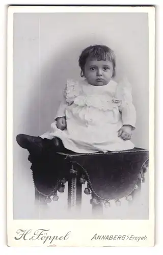 Fotografie H. Föppel, Annaberg i / Erzgeb., Portrait niedliches Kleinkind im weissen Kleid auf Tisch sitzend