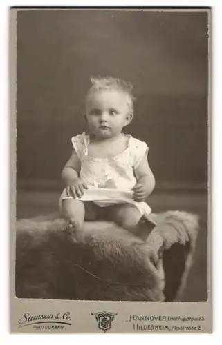 Fotografie Samson & So., Hannover, Portrait niedliches Kleinkind im weissen Hemd auf Fell sitzend
