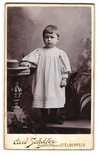 Fotografie Carl Schäfer, Elberfeld, Portrait kleines Mädchen im weissen Kleid an Tisch gelehnt