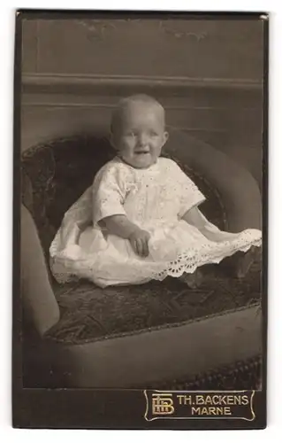 Fotografie Th. H. Backens, Marne, Portrait niedliches Kleinkind im weissen Kleid auf Sessel sitzend