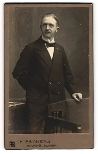 Fotografie Th. H. Backens, Marne i. Holstein, Portrait eleganter Herr mit Fliege an Stuhl gelehnt