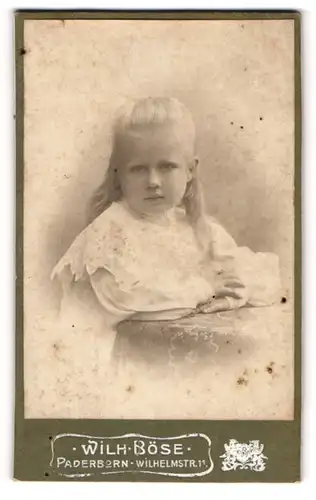 Fotografie Wilh. Böse, Paderborn, Portrait kleines Mädchen im weissen Kleid an Tisch gelehnt