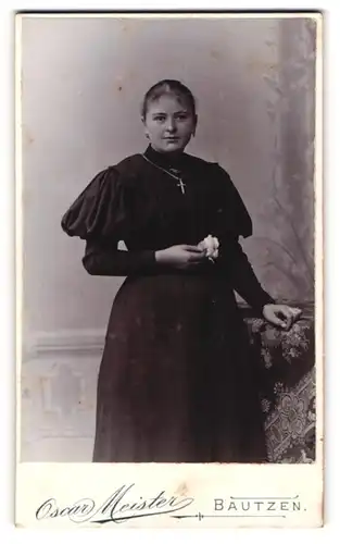 Fotografie Oscar Meister, Bautzen, Portrait junge Dame im schwarzen Kleid mit Kreuzkette und Blume