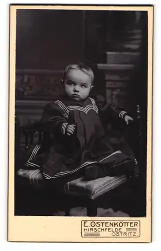 Fotografie E. Ostenkötter, Hirschfelde, Portrait niedliches Kleinkind im modischen Kleid auf Stuhl sitzend