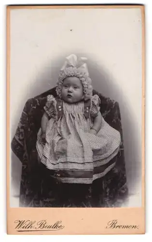 Fotografie Wilh. Beulke, Bremen, Baby im karierten Kleidchen mit Perücke sitzt auf einem Stuhl