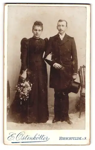 Fotografie E. Ostenkötter, Hirschfelde i. S., Ehepaar in schwarzer Garderobe mit Blumenstrauss und Hut