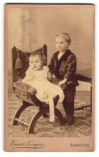 Fotografie Ernst Tremper, Hannover, Kleinkind in weissem Kleid auf Sessel mit stehendem grossen Bruder daneben